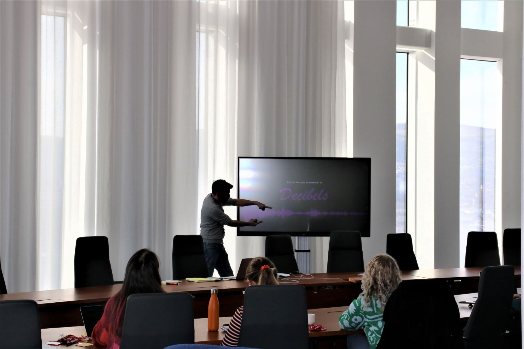 Alan Hook stands next to a television, in front of a table of students. The screen has the word