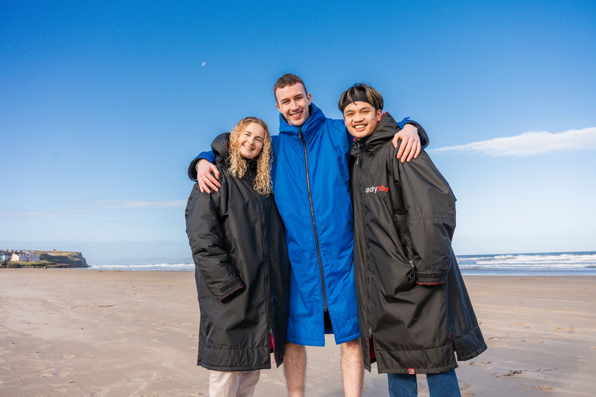 Three students standing on the beach in dry robes, arms around each other