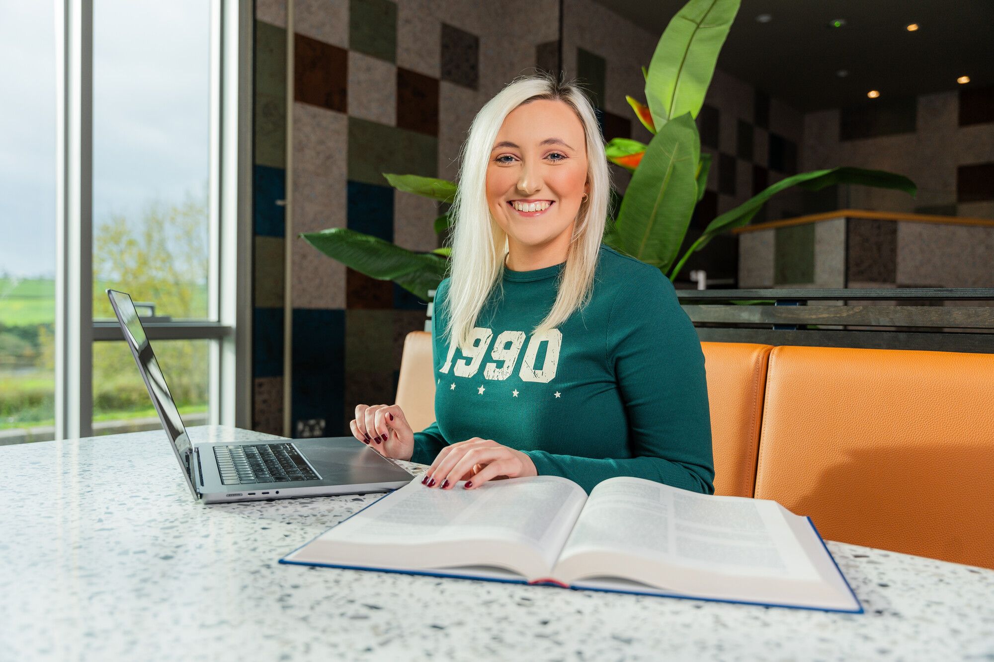 Female student with blonde hair and wearing green jumper, sitting at a canteen table with a laptop and book, smiling directly at the camera