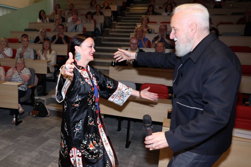 Two people at the front of a lecture theatre reach toward each other with open arms, as seated attendees clap in tiered rows behind them.