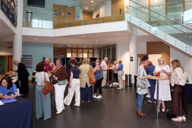 A wide view of a bright indoor foyer where multiple attendees stand around high tables, eating and chatting during a catered networking event near a staircase.