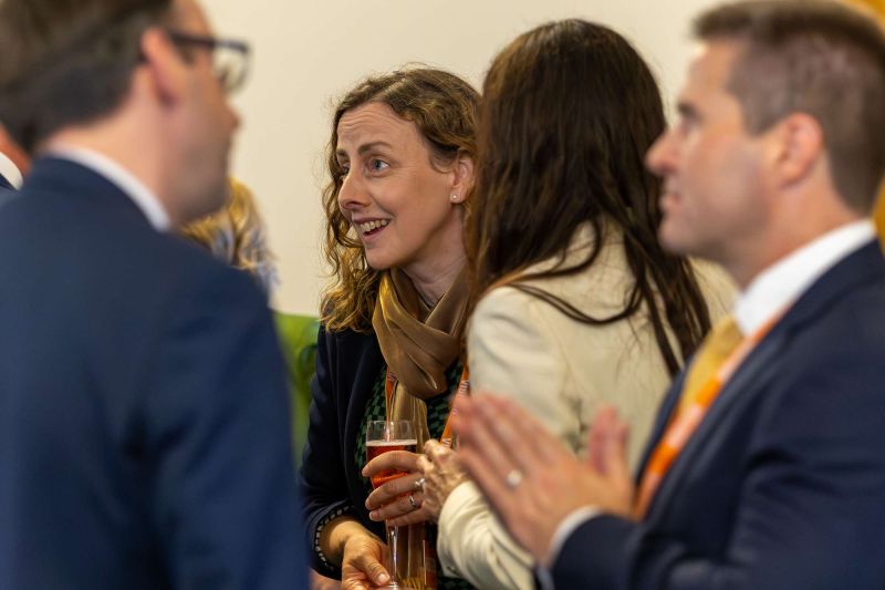Audience members seated at a formal event wearing lanyards; two individuals lean toward each other in conversation while others sit nearby in a conference-style setting.