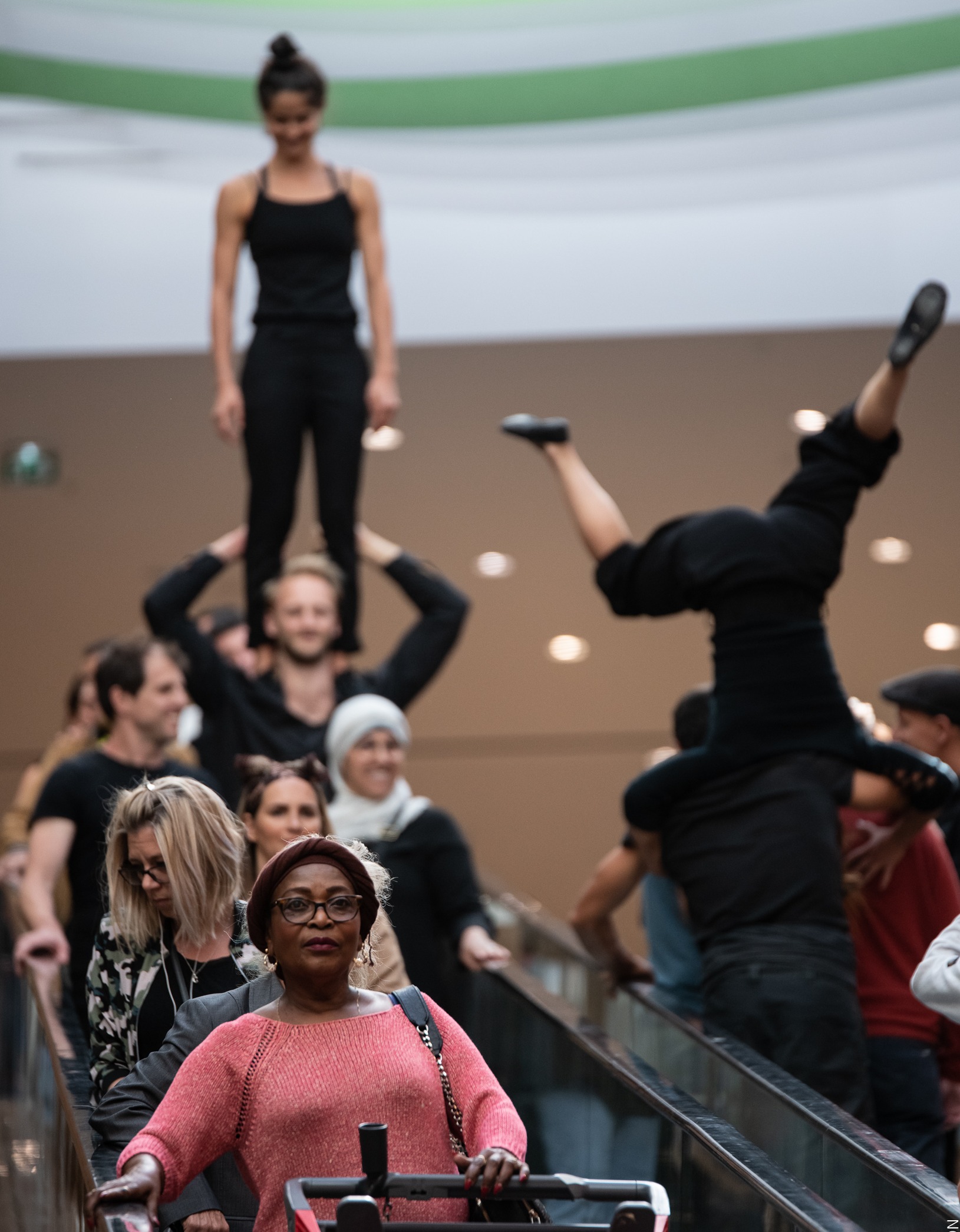 Acrobats dressed in black stand on each others shoulders and do handstands on a shopping centre escalators as others continue with their shopping.