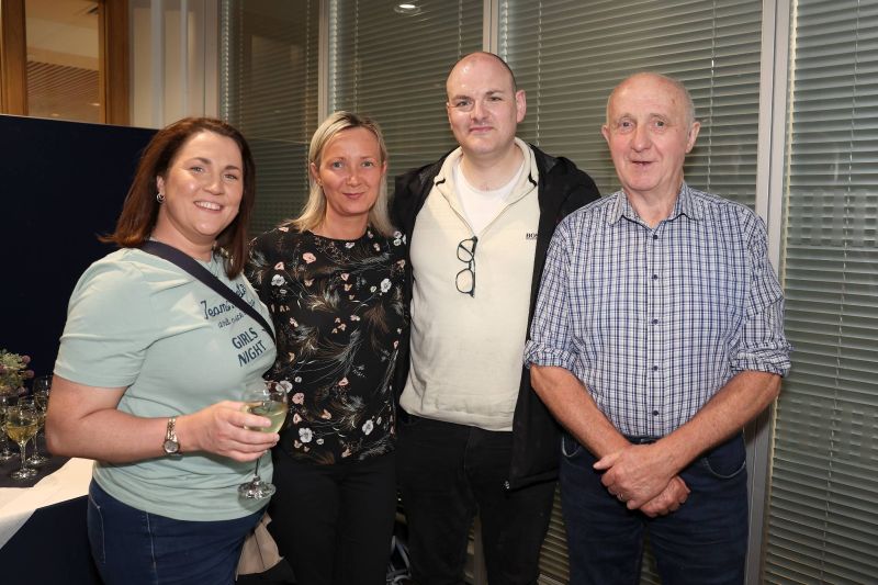 Four people stand together indoors posing for a photo, one holding a glass of white wine, with drinks and a table visible in the background.