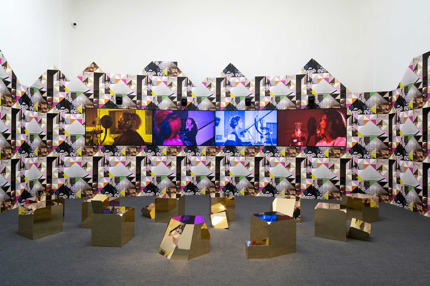 Venice Biennale 2024 artwork install by Sonya Boyce presents four widescreen TV monitors featuring four female vocalists singing at microphones in different studios. The monitors are mounted on walls covered in a kaleidscopic pattern of colourful geometric shapes and foregrounded with a number of geometric gold scultpures placed on the floor.