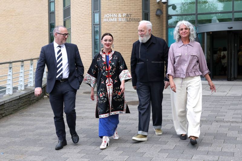 Four people walk side by side outside a brick building with a sign reading “John & Pat Hume Building”, heading toward the camera along a paved walkway.