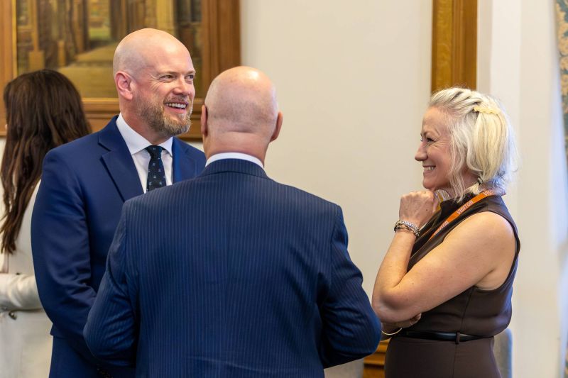 Three people stand in conversation indoors, dressed in formal attire, with framed artwork visible on the wall behind them.