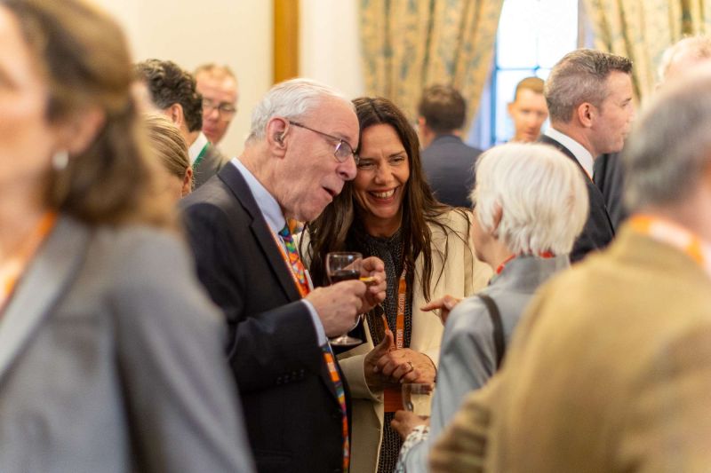Attendees wearing business attire and visitor lanyards conversing in a busy room, with artwork visible in the background.