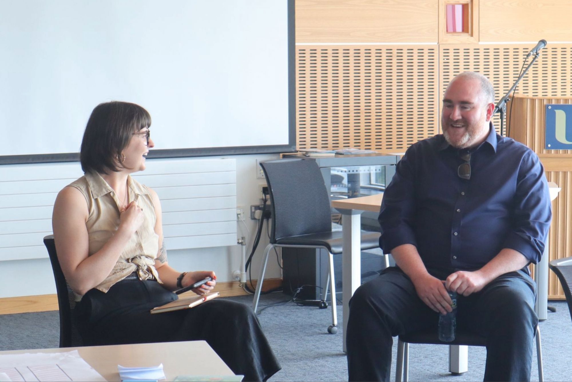 photo of a female and male sitting in chairs in a classroom. Female is interviewing male infront of an audience.