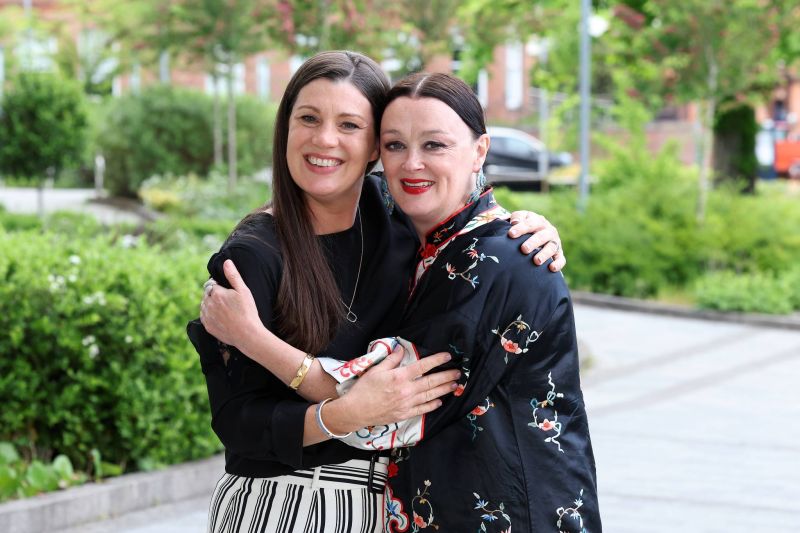 Two people stand outdoors on a path with greenery behind, posing close together with arms around each other for a photo.
