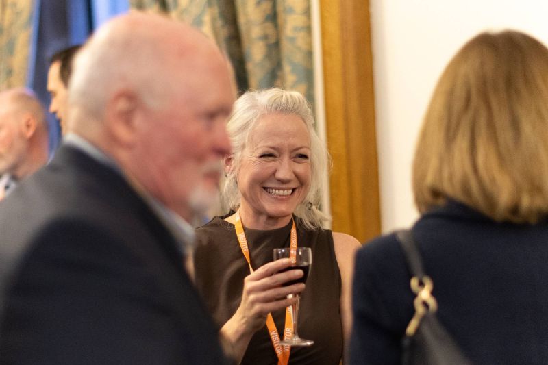 A person wearing a visitor lanyard holds a glass of red wine while talking with others in a warmly lit indoor setting during a networking event.