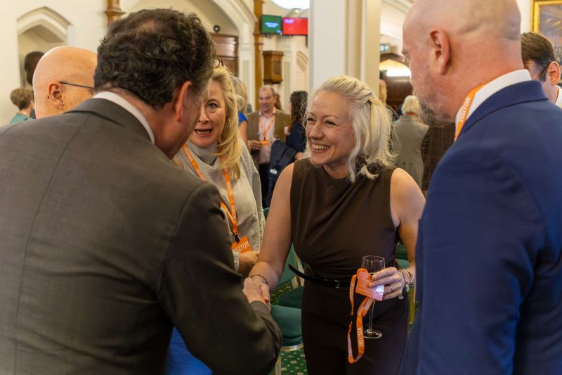 Attendees at a formal networking event greeting each other; one person shakes hands while holding a drink, with others in business attire nearby.