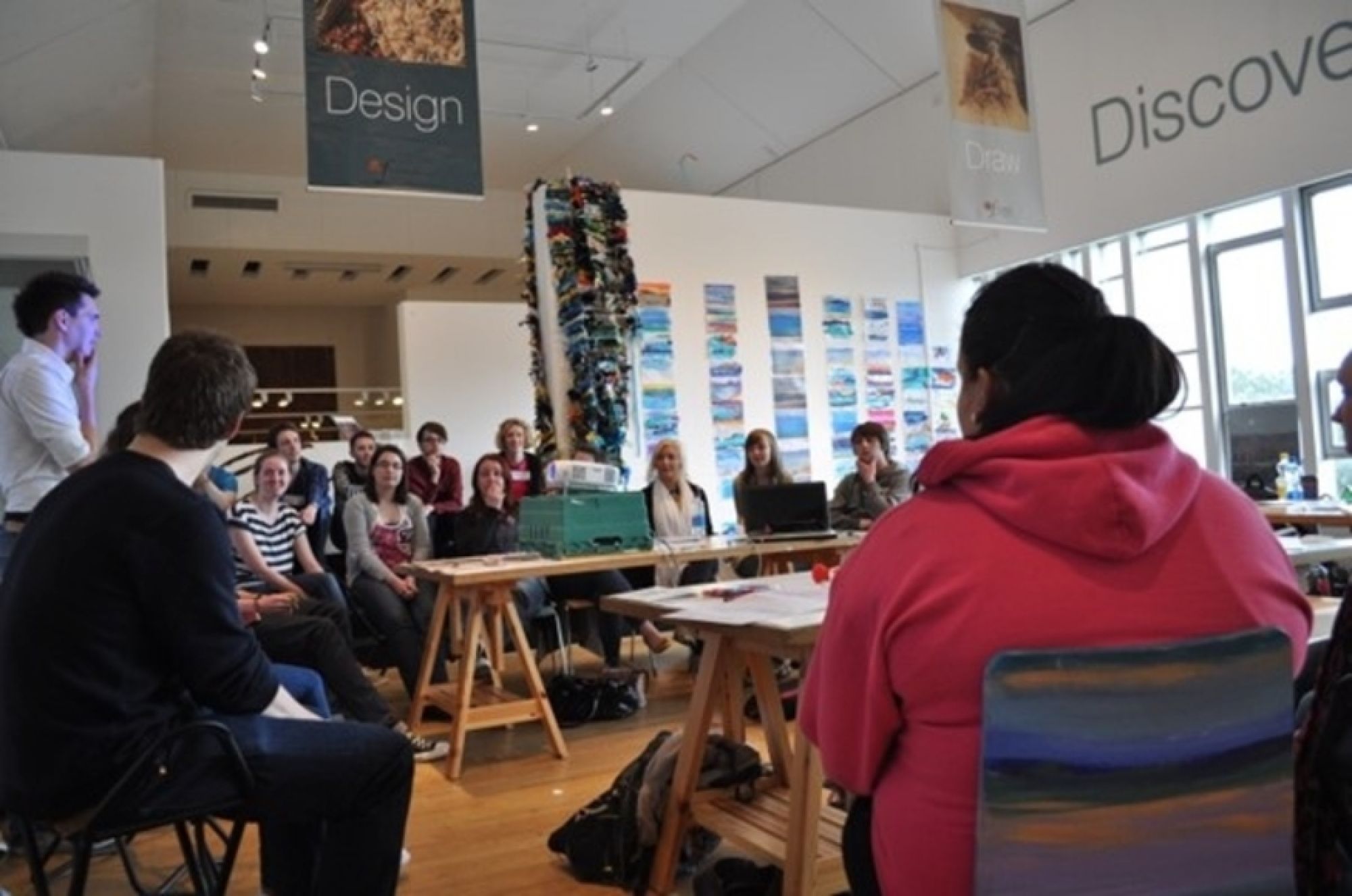 A group of students are seated around a central space, as Alan Hook delivers an introduction to a Hack Day session