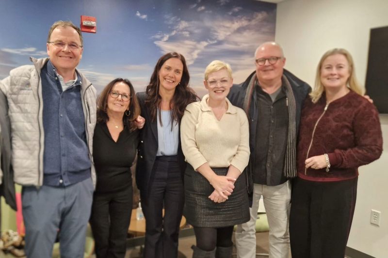 group of four women and two men standing in an office space with their arms around eachother\'s shoulders