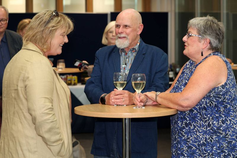Three people at an indoor event registration table, with two behind the table checking a phone and discussing details while a third person hands over a document; clipboard, pens, and glasses are on the table.