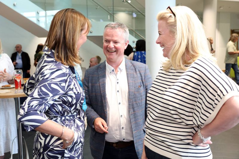 Three people stand together in a bright indoor space, chatting and laughing near a high table with drinks during a networking event.