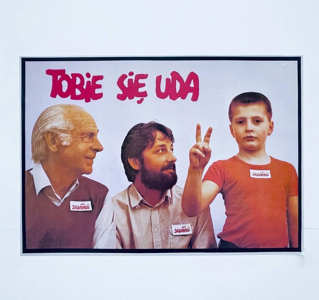 A poster image of two men look at a young boy standing, facing us, fashioning the peace sign with his fingers.