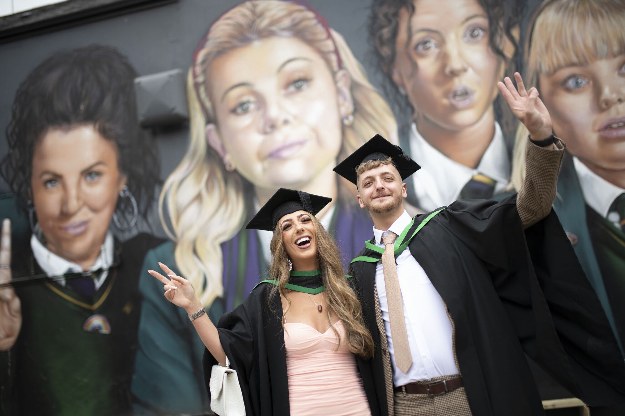 Two graduates in front of Derry Girls wall painting