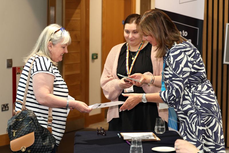 Three people at an indoor event registration table, with two behind the table checking a phone and discussing details while a third person hands over a document; clipboard, pens, and glasses are on the table.
