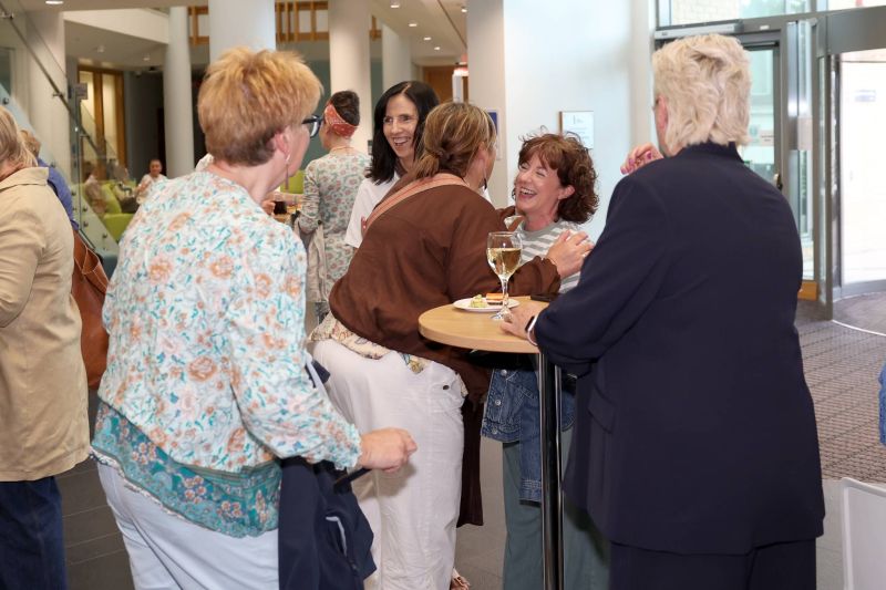 A group of attendees stand around a high table in a bright foyer, chatting and laughing while holding drinks and small plates during a networking event.