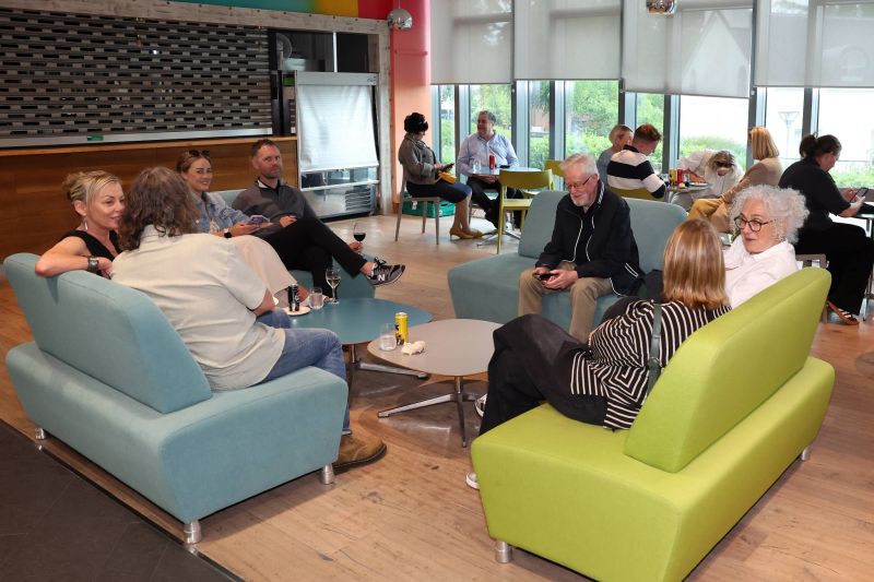 A group of people sit on sofas and chairs in a lounge-style area, talking and using phones around low tables with drinks.