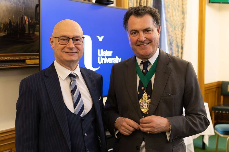 Two attendees in formal business attire standing side by side at an indoor event; one wears a ceremonial chain, with a screen behind displaying “Ulster University.”