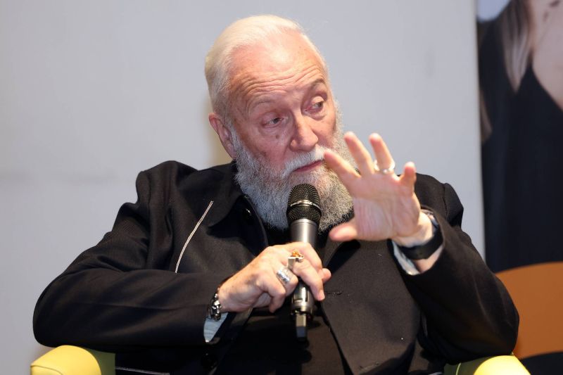 Close-up of a speaker holding a microphone, gesturing with one hand while speaking during a seated discussion or panel.