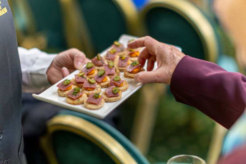 A hand selects a canapé from a serving tray holding small bite-sized snacks topped with sliced meat and garnishes, held by another person at an indoor event.