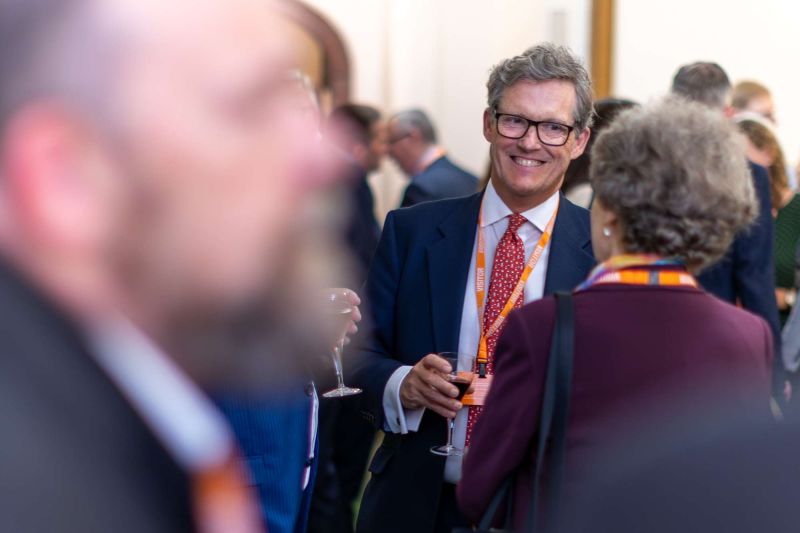 Group of event attendees in formal business attire standing closely together and conversing; one person holds a drink, with blurred figures indicating a busy networking setting.