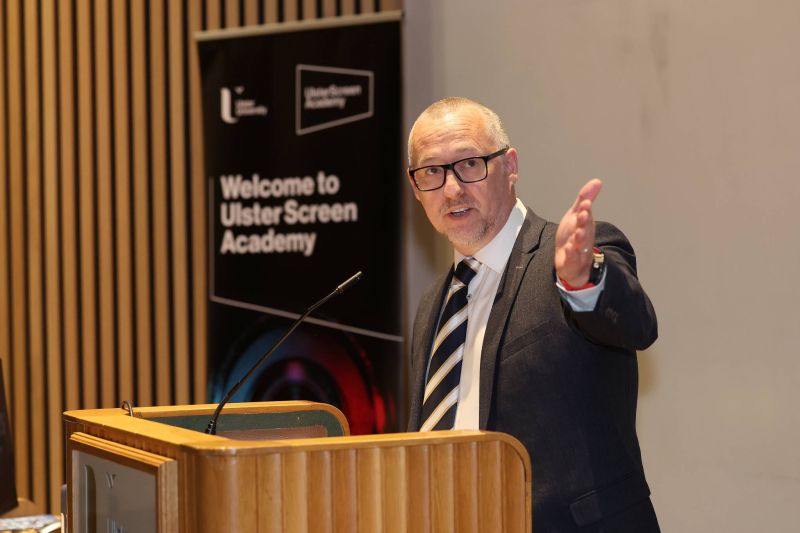 A speaker stands at a lectern with a microphone, gesturing outward during a presentation, with a banner behind reading “Welcome to Ulster Screen Academy”.